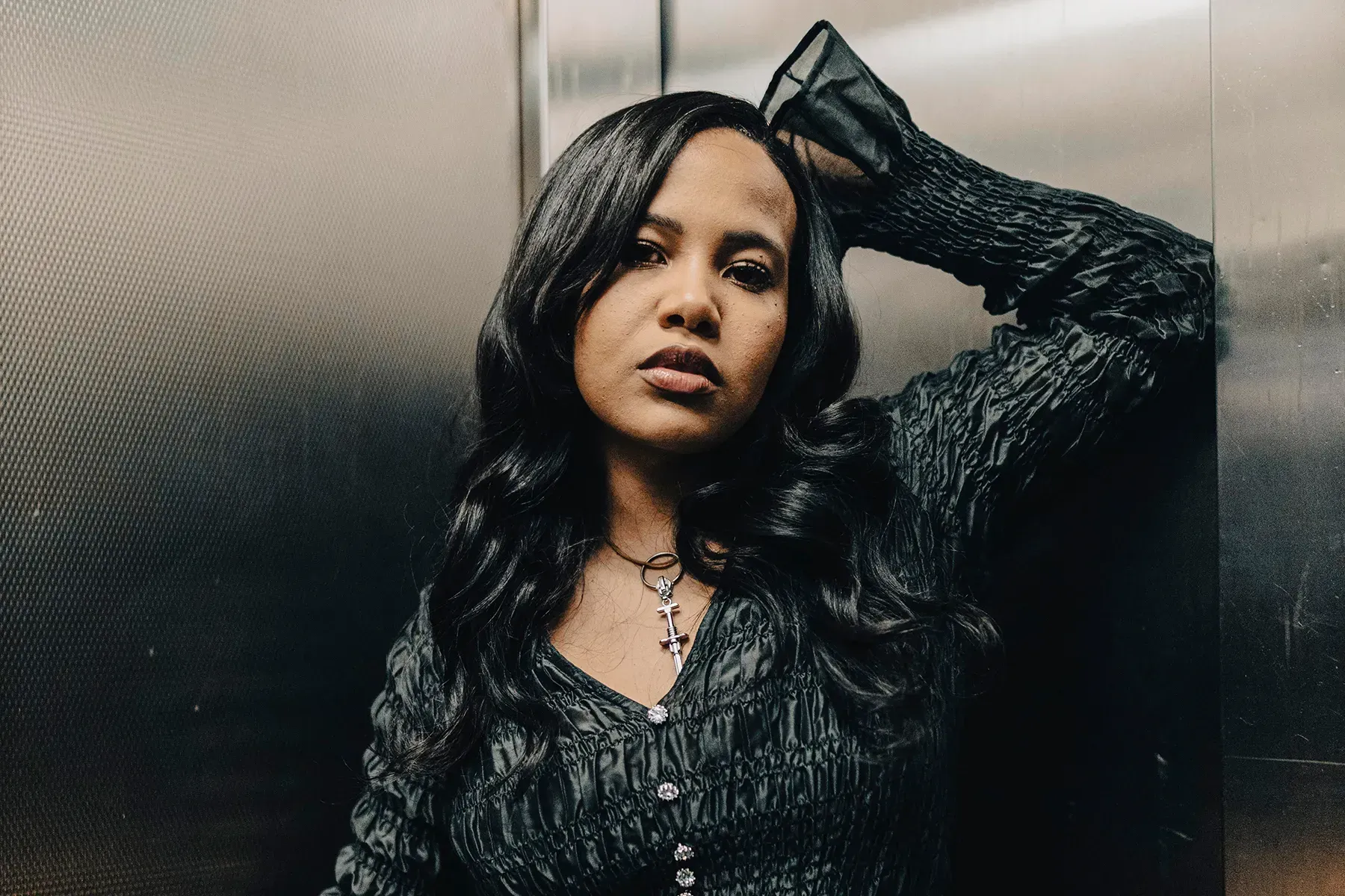 Person posing in a dark textured outfit against a metallic wall, hand behind head, looking at the camera