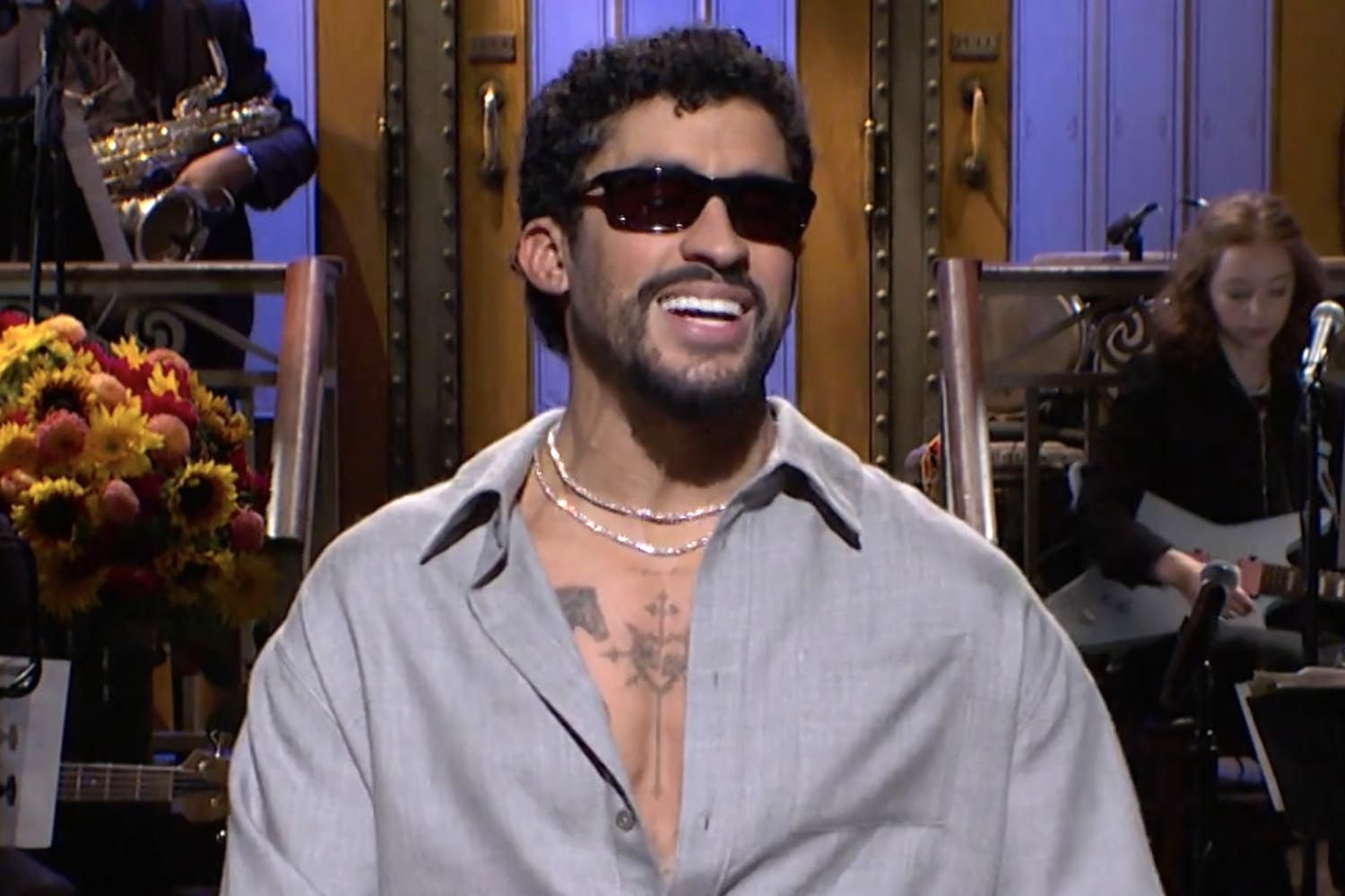 Man in sunglasses and gray shirt smiling on a late-night talk show set