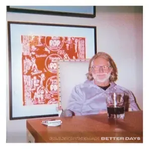 Man sitting at a desk with a drink, in front of framed artwork on a wall.