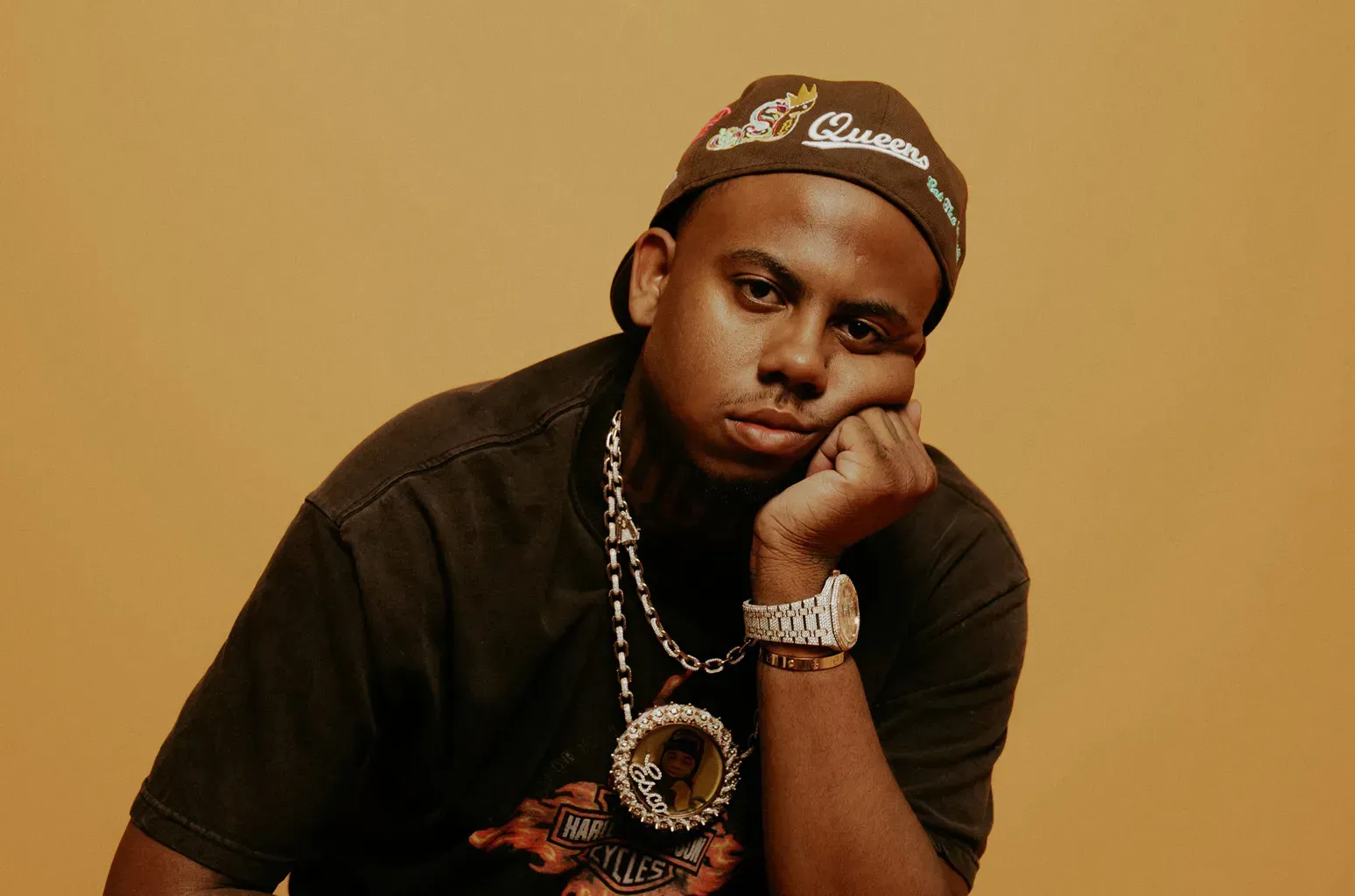Person in black clothing and cap, resting chin on hand against a warm orange background.