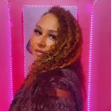 Portrait of a person with curly hair in a furry coat, posing against a pink neon-lit background