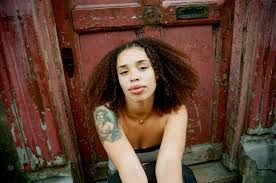 Woman seated against a weathered red wooden door, looking at the camera, with a visible arm tattoo.