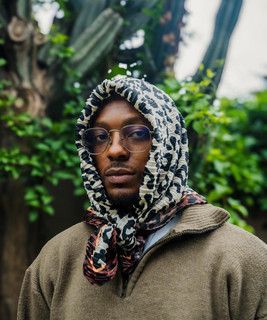 Person wearing glasses and a patterned headscarf, standing in a leafy outdoor setting.