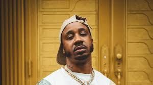 Man wearing a backward cap and gold chain standing before ornate golden doors, looking at the camera.