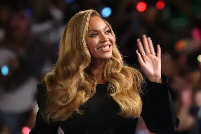Smiling woman with long blonde hair waving at an event, wearing a black outfit