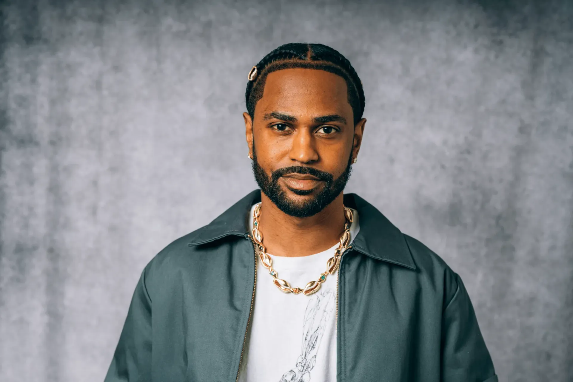 Portrait of a man with braided hair and beard, wearing a teal shirt and gold chain against a gray backdrop
