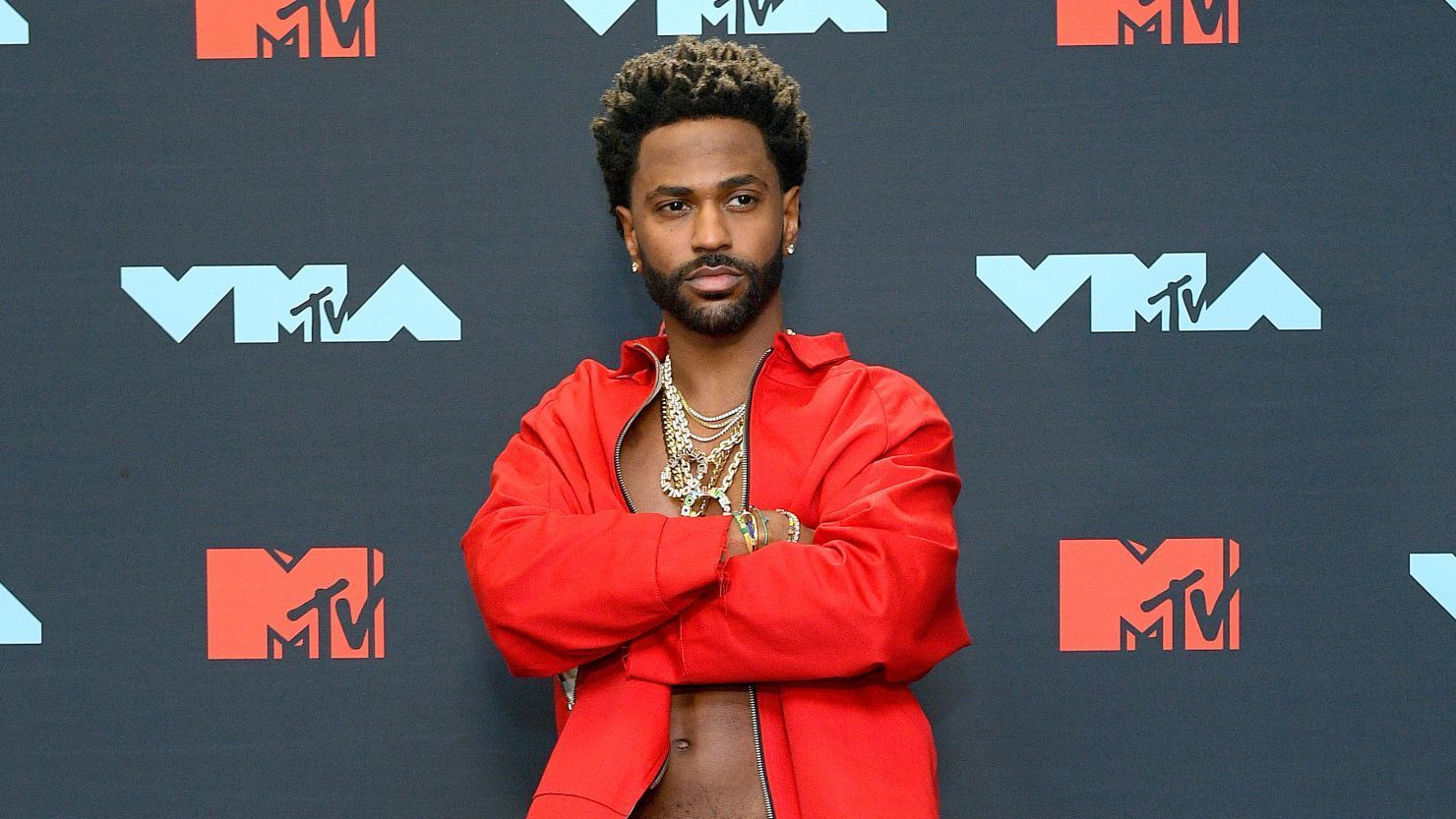 Person in a red jacket posing with arms crossed on an MTV VMA red carpet backdrop