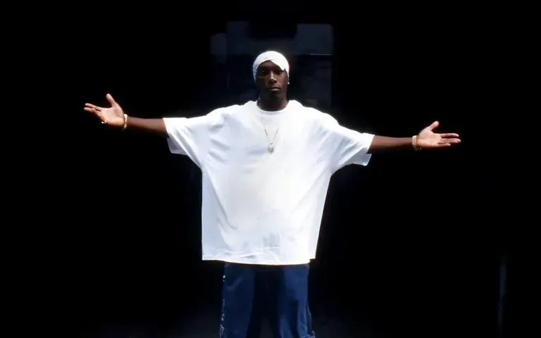 Person in a white shirt and cap stands on a dark stage with arms outstretched.