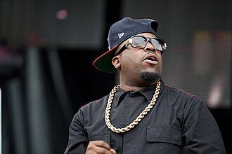Man in black shirt and cap with gold chain, looking off to the side on stage