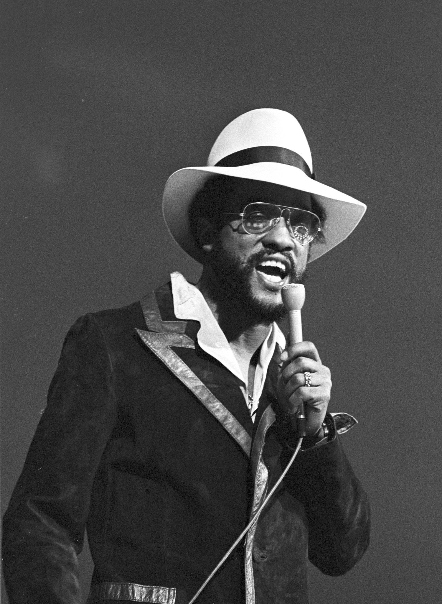 Man in a white hat and dark suit speaking into a microphone onstage