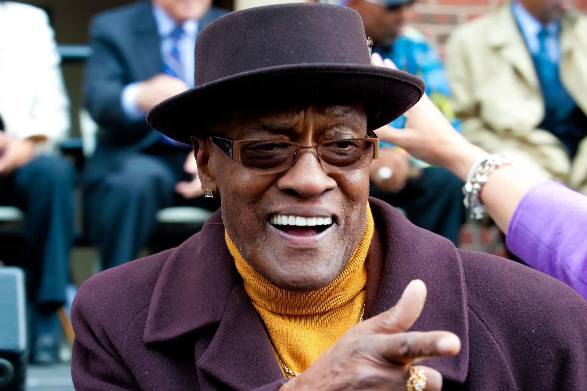 Smiling man in a hat and glasses, wearing a purple coat and yellow sweater, seated outdoors at an event