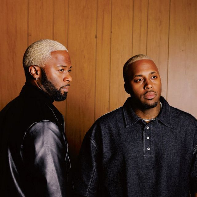Two men in dark clothing stand against a wood-paneled wall, looking off to the side.