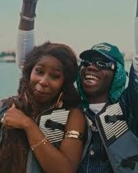 Two people smiling with arms raised outdoors, wearing sunglasses and patterned clothes.