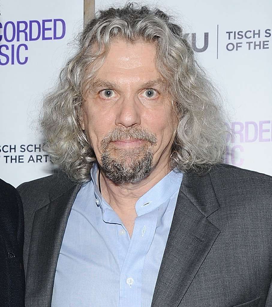 Man with curly gray hair and goatee in a gray blazer at a media event backdrop