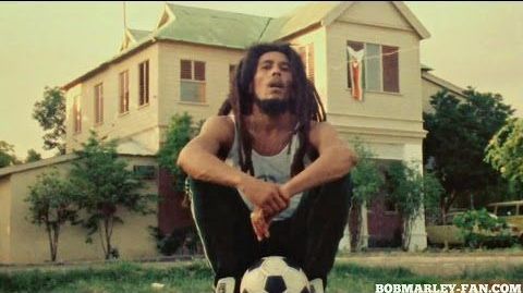 Man sitting on a soccer ball in front of a house, with his arms resting on his knees.