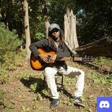 Person sitting outdoors playing a sunburst guitar in a wooded area, wearing a cap and sunglasses.