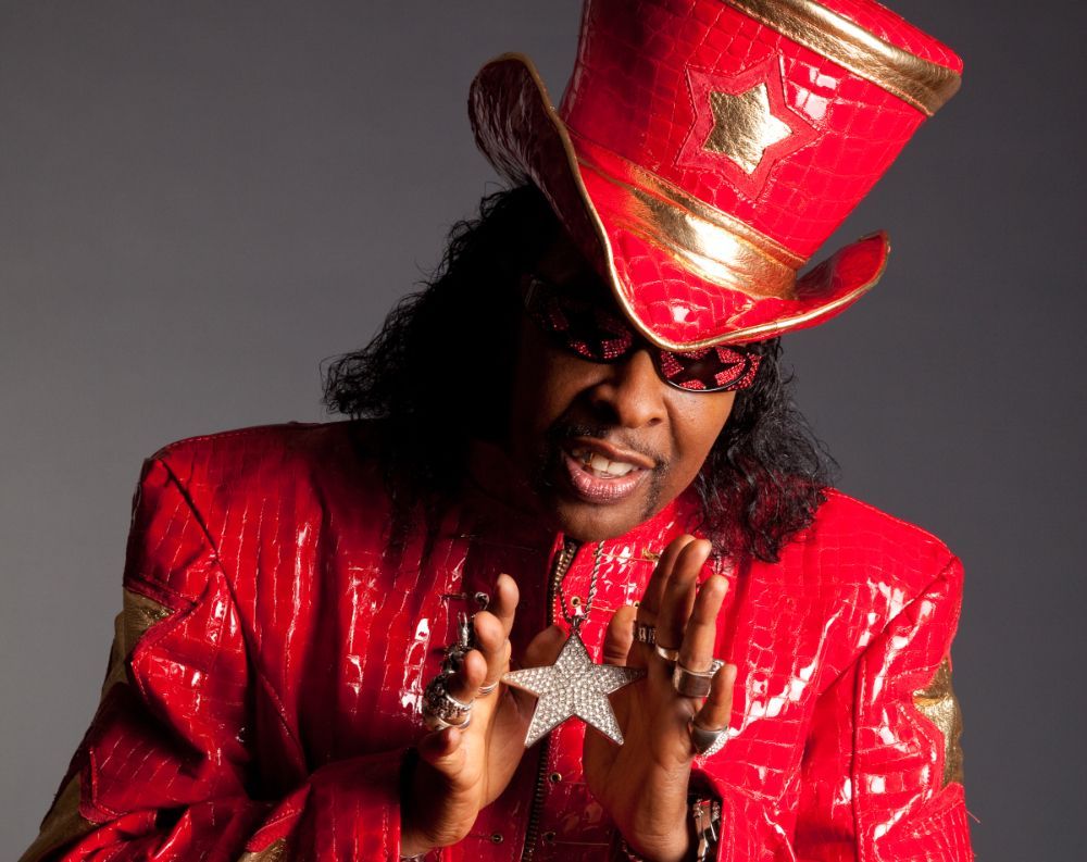 Person in a shiny red suit and tall hat posing against a gray background, hands raised with a star pendant visible