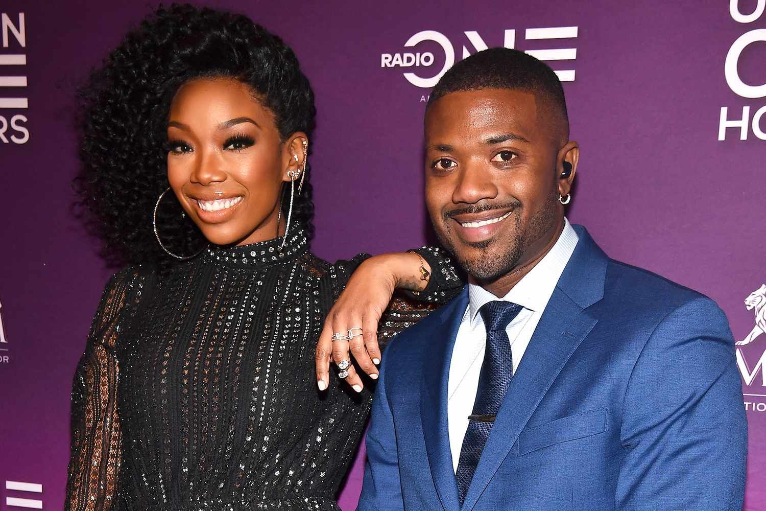 Two people smiling at a red carpet event with a purple backdrop, one in a black dress and one in a blue suit