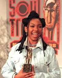 Smiling performer with braided hair and chains on stage, wearing a white shirt against a pink-purple background