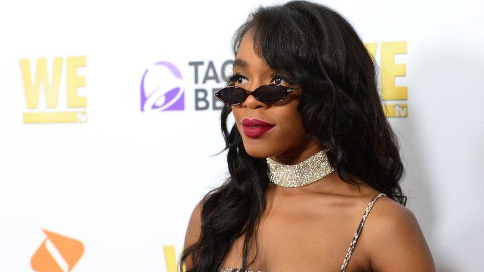 Woman in black sunglasses and sparkly choker at a red-carpet event backdrop