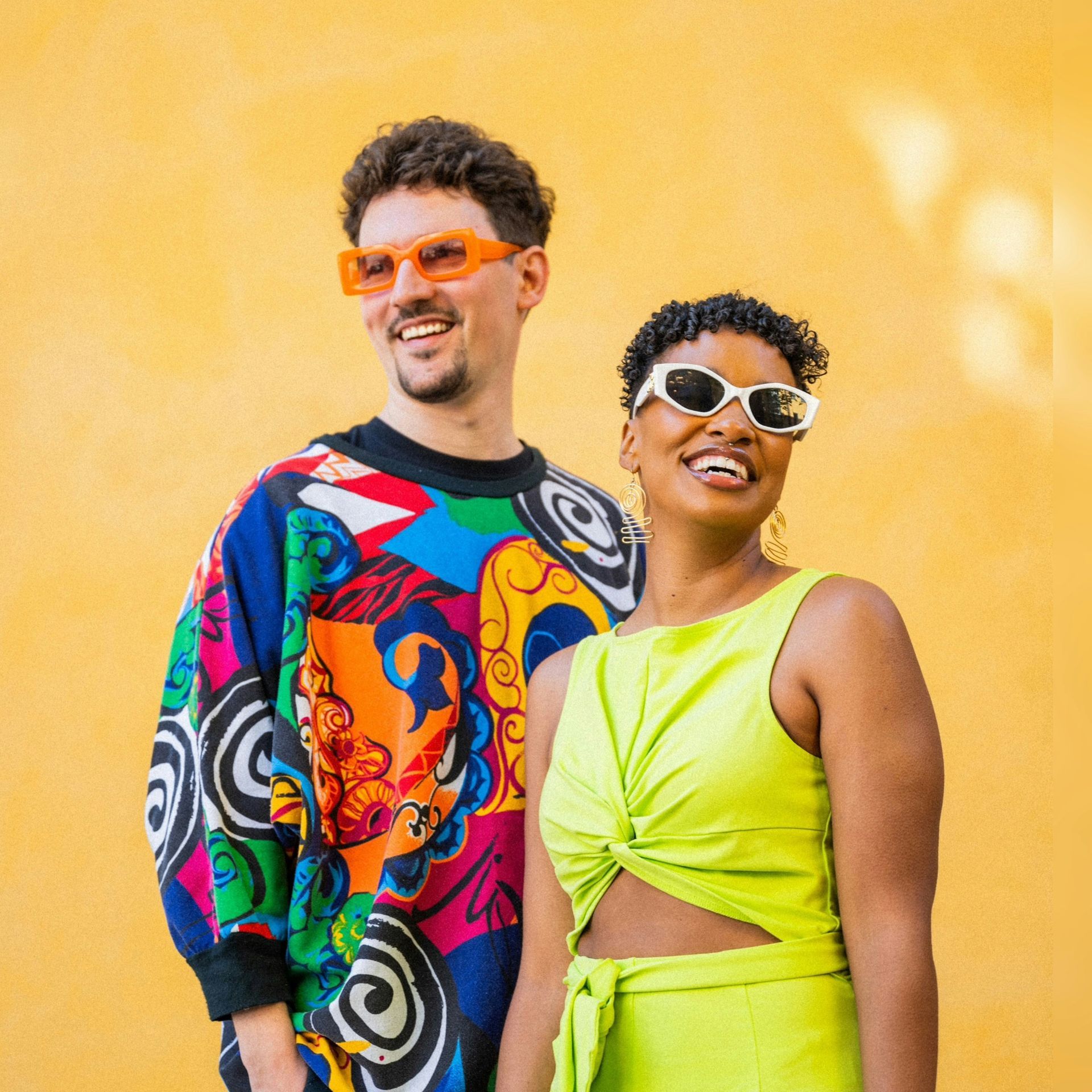 Two smiling people in colorful clothes and sunglasses posing against a yellow wall
