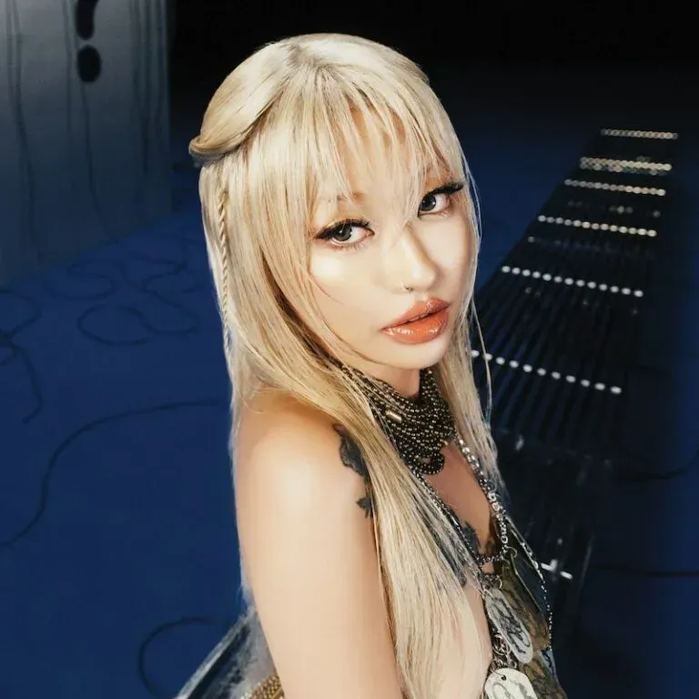 Blonde person with long hair and dramatic makeup, wearing a black outfit, posing in a dark blue-lit room.