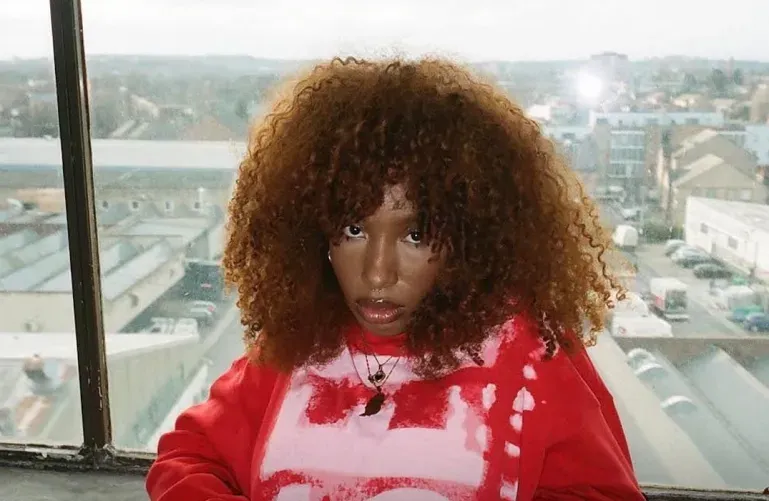 Person with curly hair in a red sweatshirt, sitting by a window with a city view.