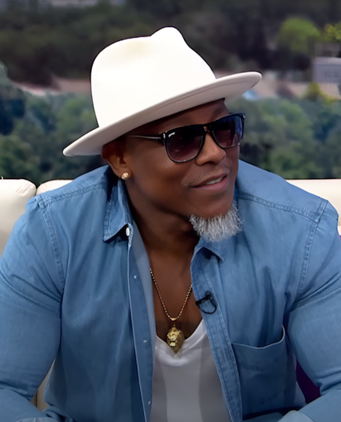 Man in white hat and sunglasses wearing a blue shirt, seated outdoors against a blurred green background