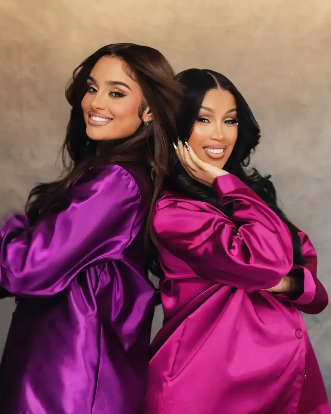 Two women in shiny purple outfits pose back-to-back, smiling against a warm studio backdrop.