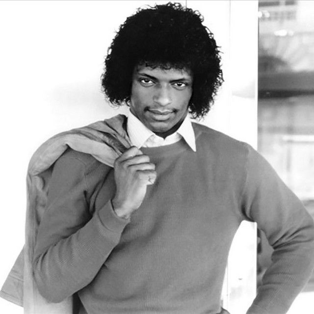 Person in a sweater with a jacket over one shoulder, standing and looking at the camera.