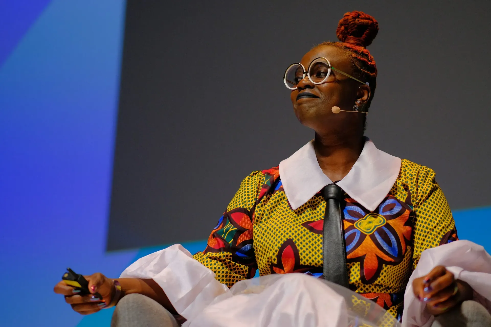 Speaker on stage wearing colorful patterned outfit and glasses, seated against a blue-lit background