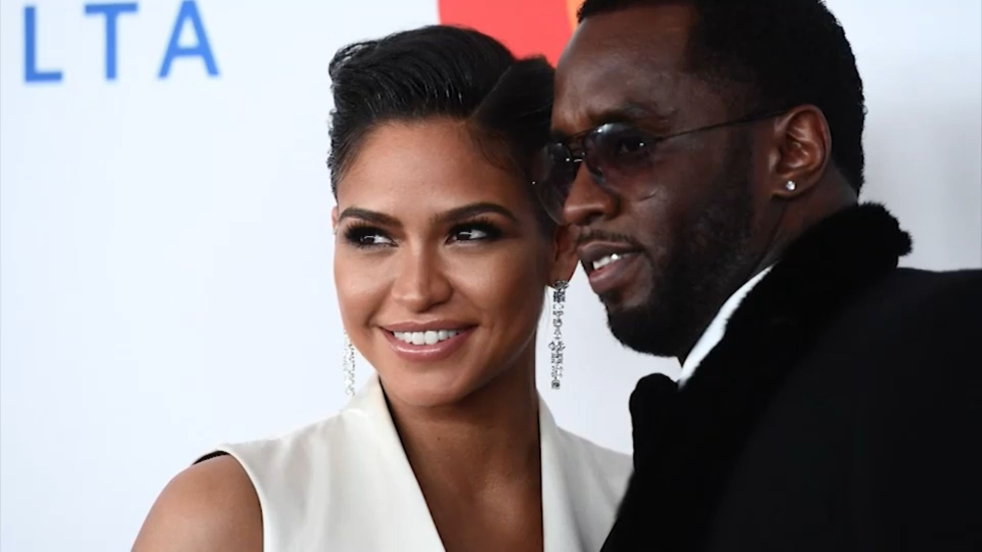 Two people smiling at a red carpet event, posing close together before a branded backdrop.