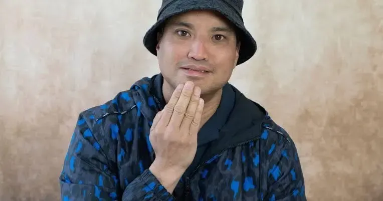 Person in a blue jacket and black hat, hand raised to chin, against a beige background.
