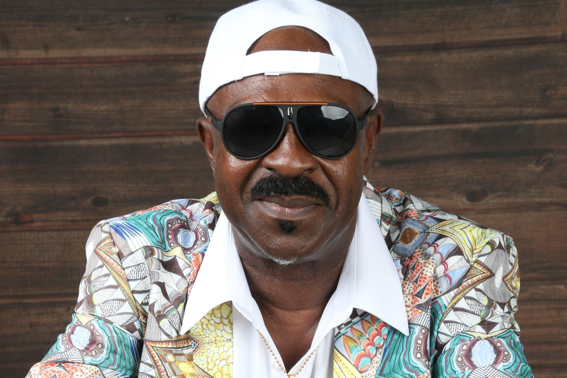 Man in sunglasses and a white cap, wearing a colorful patterned shirt, seated against a wooden wall.