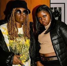 Two people posing indoors, wearing dark jackets and casual outfits, with framed art on the wall behind them