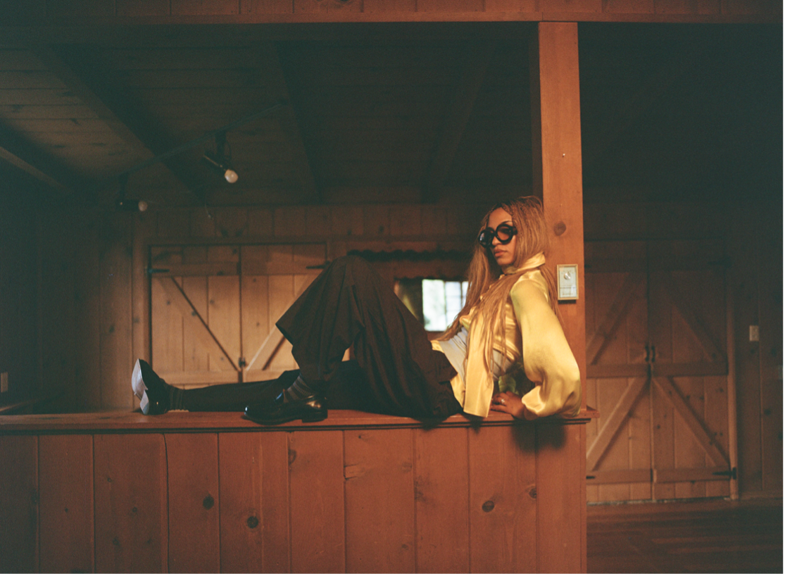 Person in black pants and yellow jacket lounges on a wooden balcony in a rustic barn-like interior.