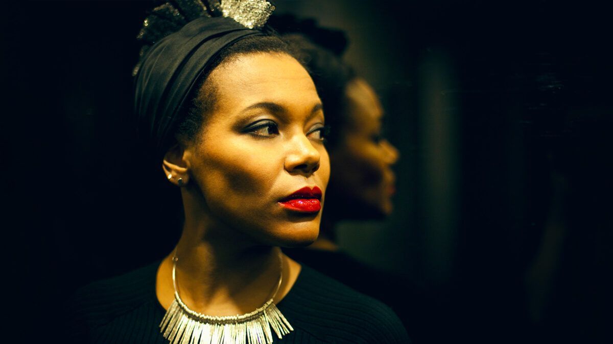 Portrait of a woman in profile, wearing a black headwrap, gold necklace, and red lipstick against a dark background