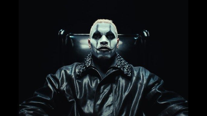 Person in a dark leather jacket, face painted white and black, seated in a dimly lit setting.