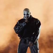 A man in a black jacket and jeans stands against an orange smoky background, hand near his mouth.