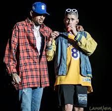 Two men posing on a dark background, one in a red plaid shirt and blue cap, the other in a yellow jersey and sunglasses