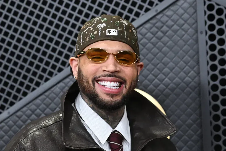 Smiling man in a leather jacket, tie, sunglasses, and patterned cap against a dark textured wall