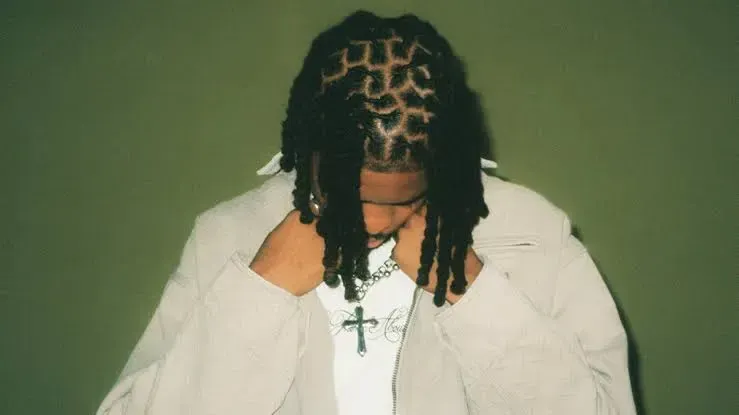 Person with braided hair in a white jacket and cross necklace against a green background