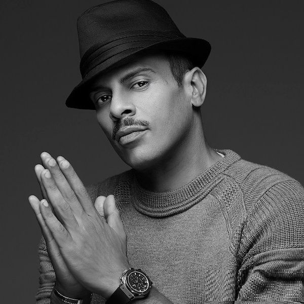 Man in a dark hat and sweater, hands clasped, posing against a plain gray background.