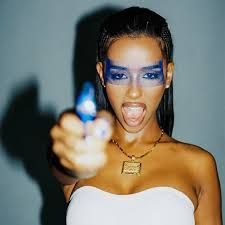 Woman in white strapless top aiming a blue toy gun toward the camera, mouth open against a dark background