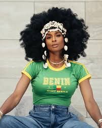 Person with large afro, green “BENIN” T-shirt, and jeans sitting against a tiled wall