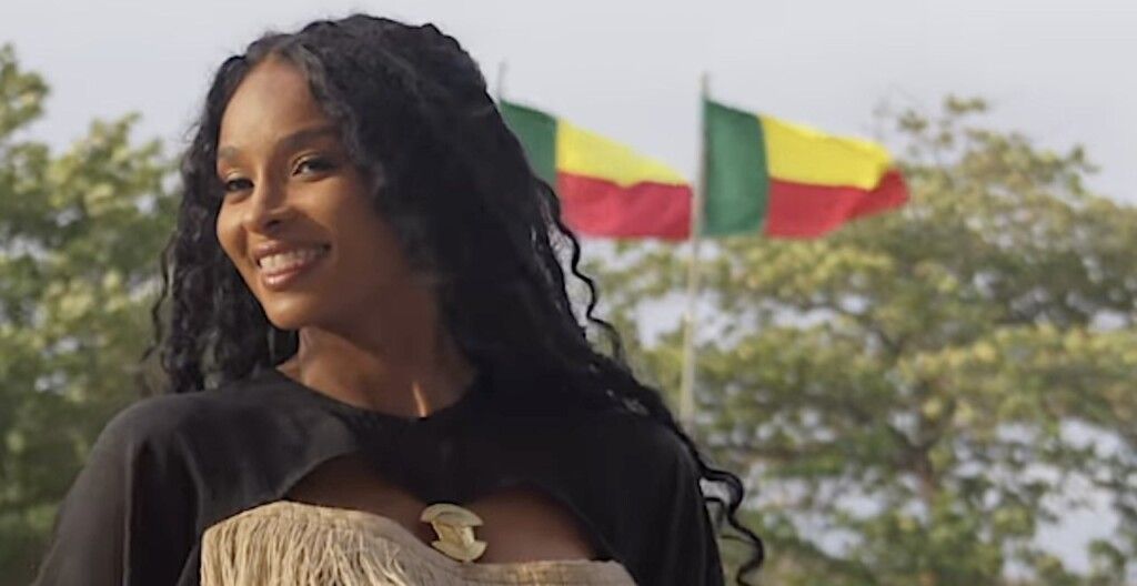 Smiling woman in black and beige dress standing outdoors with red, yellow, and green flags behind her