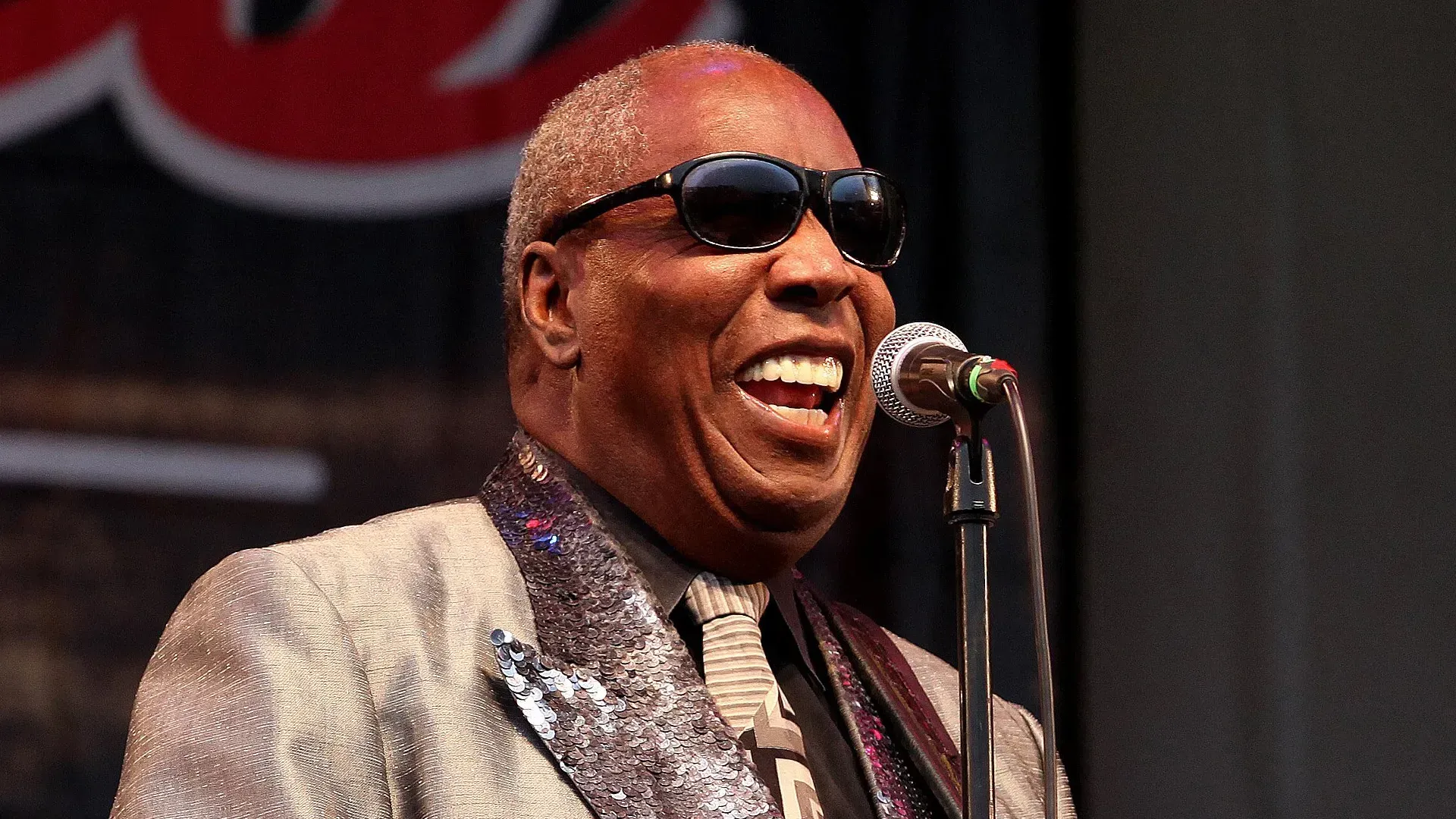 Musician in sunglasses singing into a microphone on stage, wearing a silver jacket.