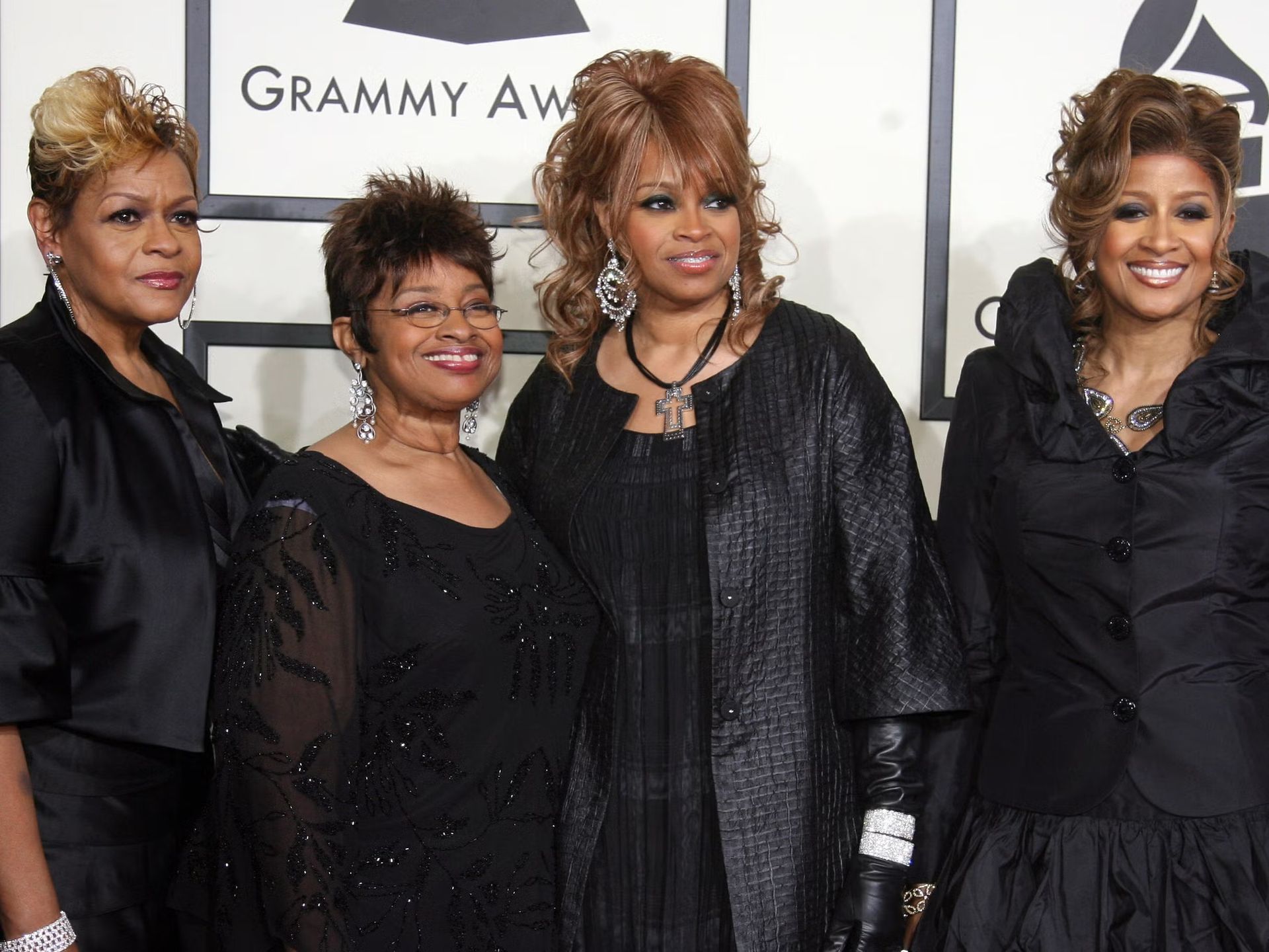 Four women posing on a Grammy Awards red carpet in black dresses and dark outfits.