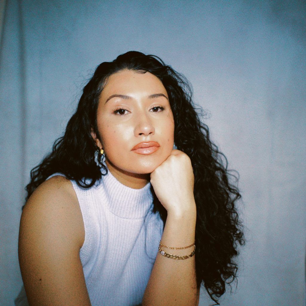 Portrait of a woman with curly hair in a sleeveless white top, resting her chin on her hand against a blue background