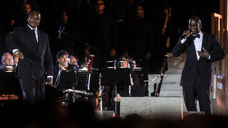 Two men in tuxedos perform with an orchestra on a dimly lit stage.